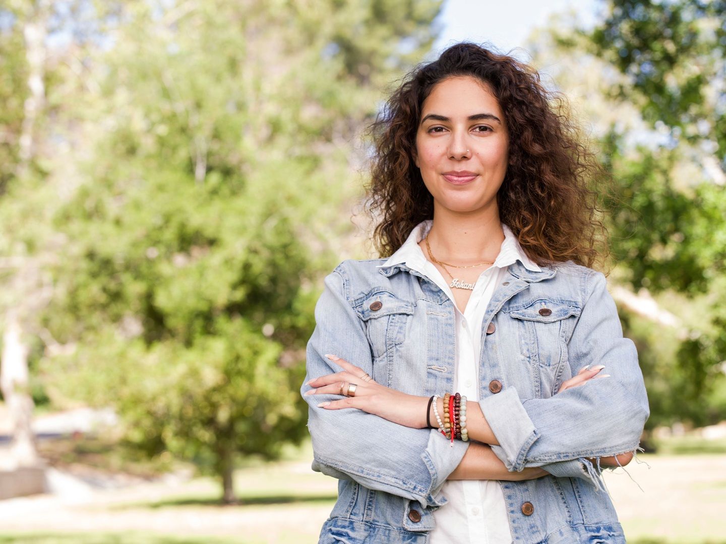 Una joven con cabello largo rizado y de brazos cruzados que tiene esclerosis múltiple sonríe al aire libre.