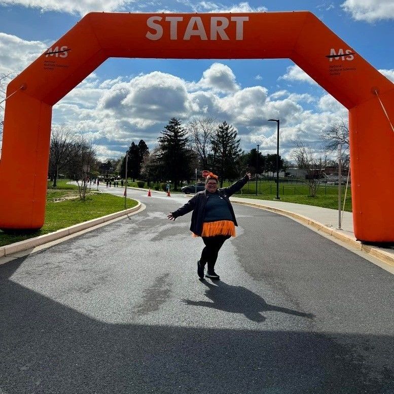 A woman with an orange bow in her head and an orange skirt poses under the orange starting line arch at a Walk MS event.