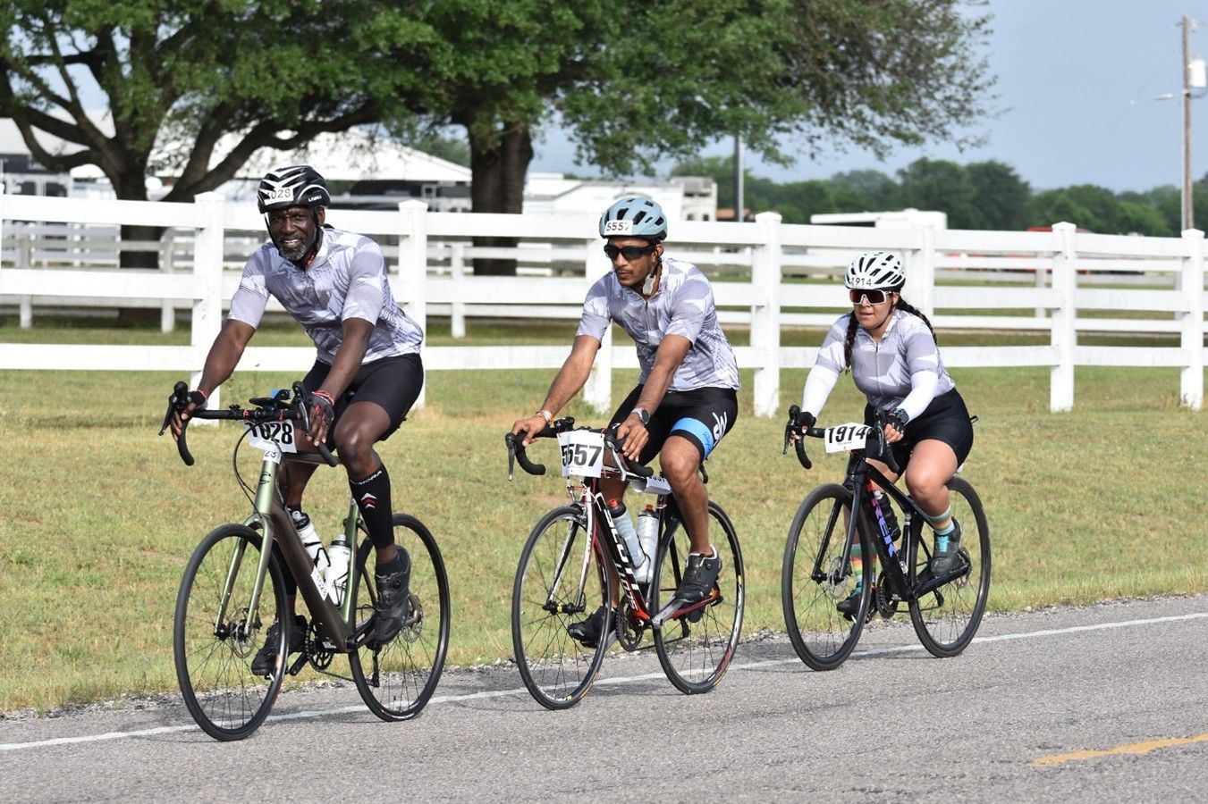 Three cyclists ride side by side during an organized event, all wearing matching gray and white cycling jerseys.