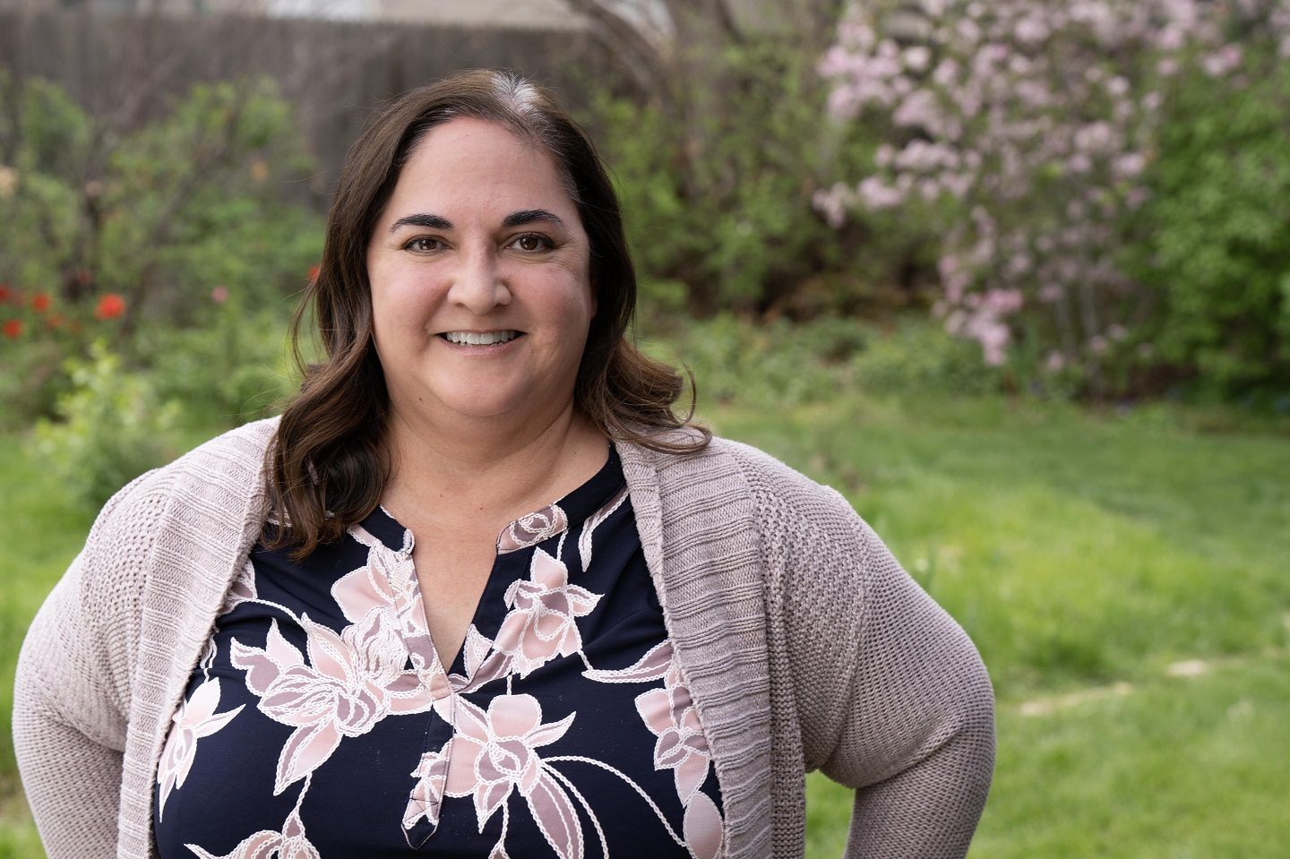 Una mujer en blusa negra floreada y suéter sonríe al aire libre.