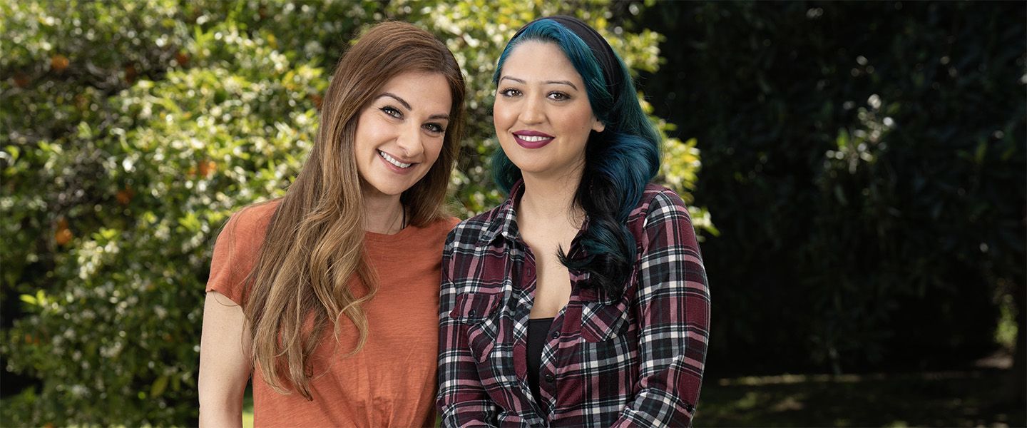 A young woman with long, black hair streaked with blue who lives with MS stands holding a cane next to another woman her age.