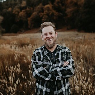 A young man with a beard wearing a plaid shirt stands in the middle of a wheat field smiling.