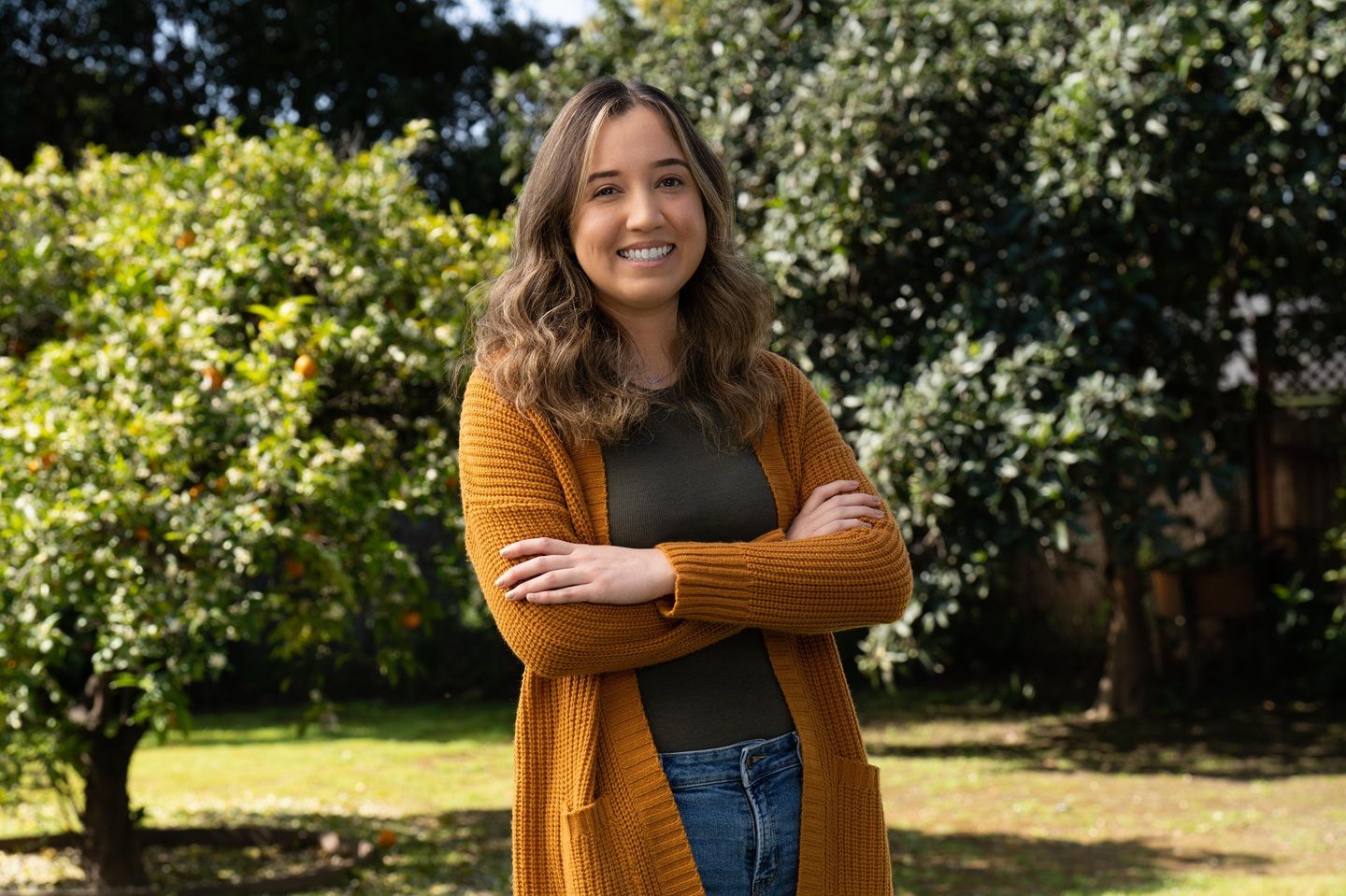Jackie, a woman with MS, smiling in an orange sweater while standing in a lawn with trees.