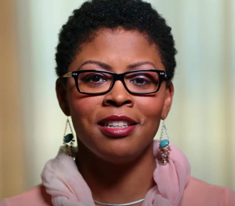A person with short brown hair wearing a pink scarf and sweater smiles at the camera.