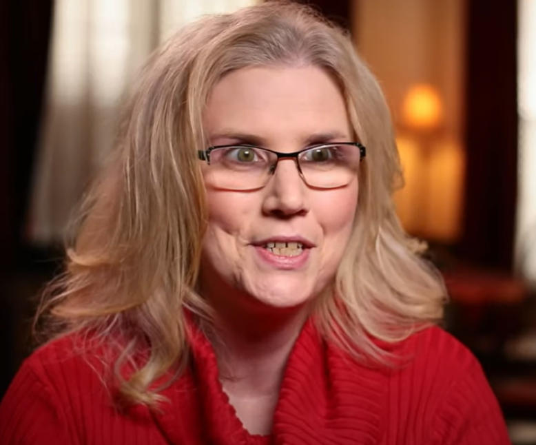A person with shoulder-length blonde hair wearing a red shirt smiles at the camera.